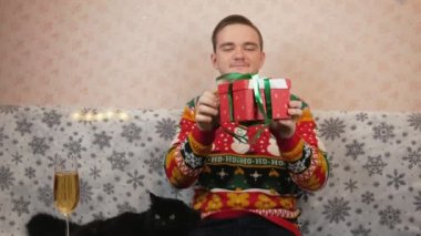 Yeni yıl süveteri giyen bir adam kanepeye oturur ve kırmızı bir kutuya hediye koyar, oyuncak ve tıbbi bir maske takar, bir bardak şampanyanın yanında, hediyeyi hangi kurdeleyle paketleyeceğini seçer, kırmızı mı yeşil mi?