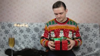 Yeni yıl süveteri giyen bir adam kanepeye oturur ve kırmızı bir kutuya hediye koyar, oyuncak ve tıbbi bir maske takar, bir bardak şampanyanın yanında, hediyeyi hangi kurdeleyle paketleyeceğini seçer, kırmızı mı yeşil mi?