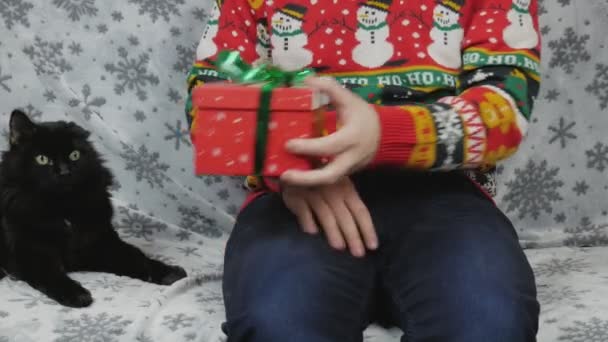 一个穿着新年毛衣的男人坐在沙发上 用红色的盒子包装礼物 包装玩具和医疗用的红色面具 旁边是一杯香槟 他选择用红色或绿色的带子包装礼物 — 图库视频影像