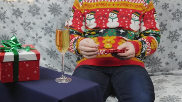 一个穿着新年毛衣的男人坐在沙发上 用红色的盒子包装礼物 包装玩具和医疗用的红色面具 旁边是一杯香槟 他选择用红色或绿色的带子包装礼物 — 图库视频影像