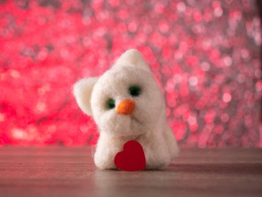 Beyaz oyuncak kedi, pembe arka planda kırmızı bir kalple masada duruyor.