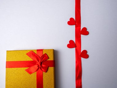 Beyaz parlak bir arka planda dört kalple çevrili uzun kırmızı bir kurdele ve kırmızı kurdeleli sarı bir hediyenin yanında uzanır.