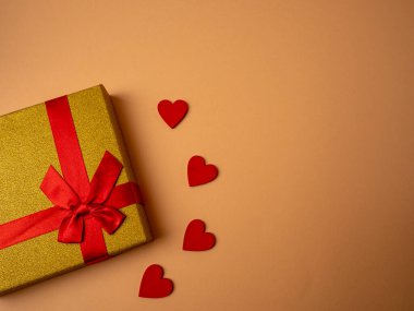 Kelebek düğümü şeklinde kurdeleli sarı bir hediye turuncu arka planda dört kırmızı kalbin yanında yatıyor.