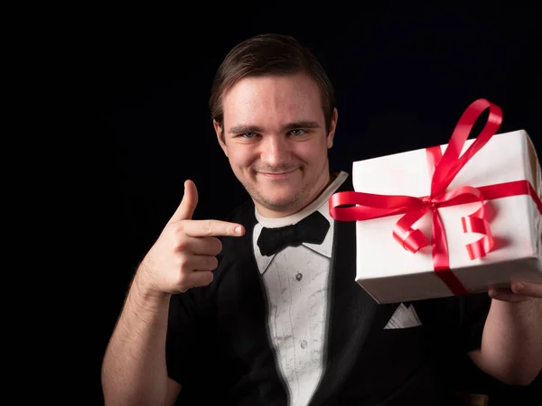 Siyah tişörtlü genç bir adam siyah arka planda beyaz bir hediyeye parmağını gösteriyor.