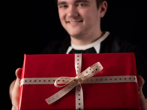 young guy in black t-shirt suit stretches out his hand with red gift on a black background