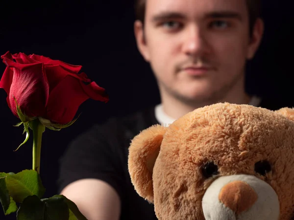 Siyah tişörtlü genç adam elinde kırmızı bir gül ve oyuncak ayı tutuyor ve siyah bir arka planda görünüyor.