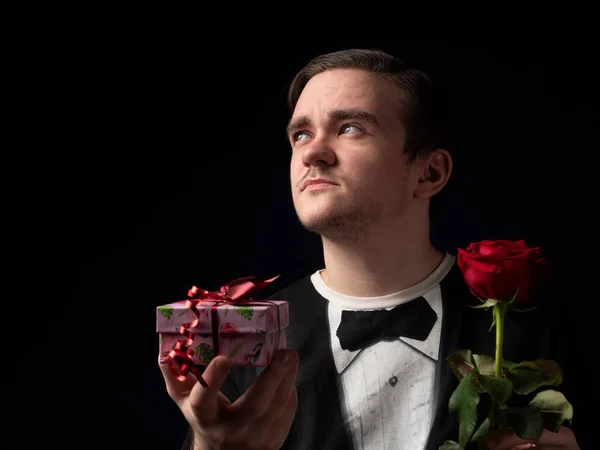 Young guy in a black t-shirt suit holding a red rose and a pink gift in his hands looking up on a black background