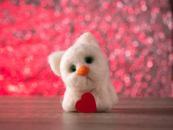 Beyaz oyuncak kedi, pembe arka planda kırmızı bir kalple masada duruyor.