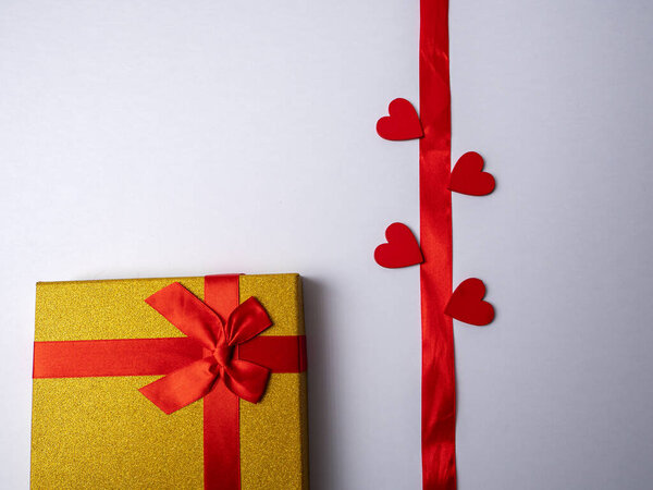 On a white bright background lies a long red ribbon surrounded by four hearts and next to a yellow gift with a red ribbon