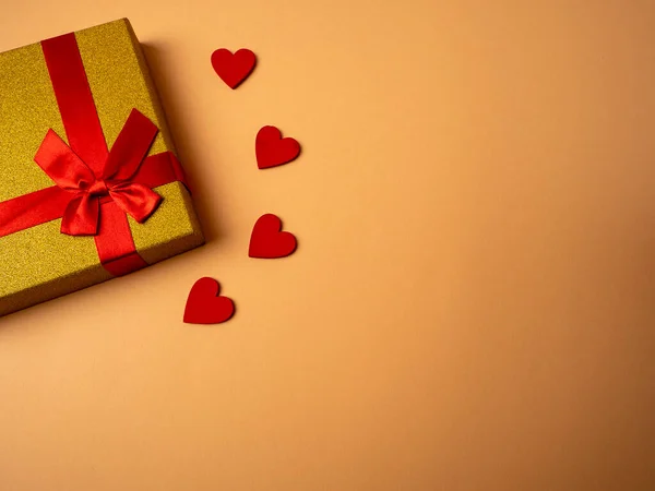 Kelebek düğümü şeklinde kurdeleli sarı bir hediye turuncu arka planda dört kırmızı kalbin yanında yatıyor.