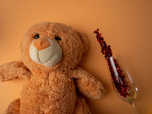 A large brown teddy bear lies next to a glass with sparkles on an orange background