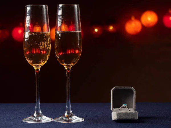 İki kadeh şampanya ve yanında beyaz bir kutu var. Üzerinde siyah bir yüzük ve ışık var.