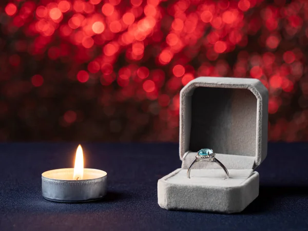 Yanan beyaz bir mum ve yanında pembe arka planda mavi taşlı bir yüzük olan bir kutu var.