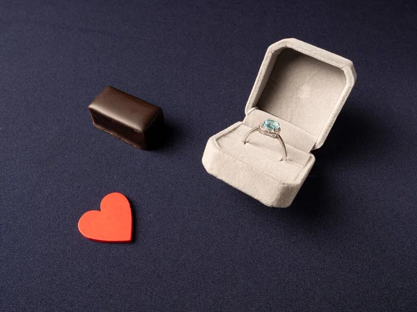 Mavi taşlı, kırmızı kalpli ve çikolatalı bir yüzüğün mavi arka planda yattığı beyaz kutu.