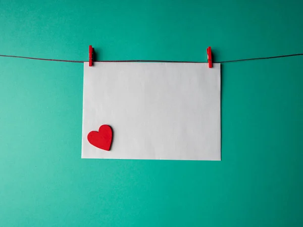 Beyaz kağıt kırmızı bir ipe bağlıdır ve iki mandalla sabitlenir ve yeşil arka planda kırmızı bir kalbi vardır. Sevgililer Günü projen için bir şablon.