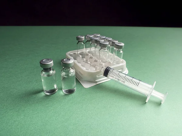İçinde aşı olan sıvının otomatik sıvı dağıtıcının tepsisinde olduğu cam şişeler ve üzerinde bir şırınga var. Cam şişeler ve şırıngalar yeşil ve siyah arkaplanda.
