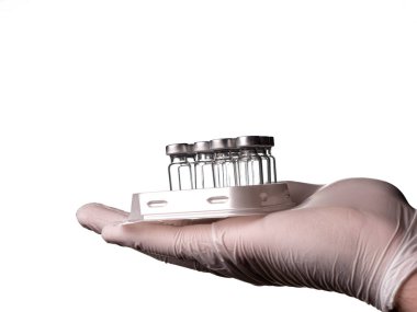 Beyaz eldivenli bir doktor, cam şişeleri ve aşı sıvısını otomatik sıvı dağıtıcının tepsisinde tutuyor. Cam şişeler ve el beyaz arka planda izole