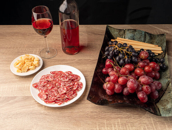 Sliced salami and cheese with rose wine, two types grapes, and Italian grissini on a tray, and on a wooden table, over black background. Top view flat lay