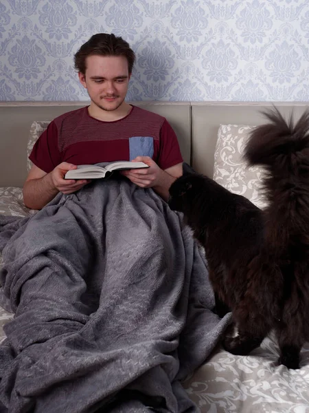 Kırmızı tişörtlü genç bir adam açık renkli bir yatakta oturur ve kalın bir kitap okur. Çalışma gününden sonra dinlenme konsepti.