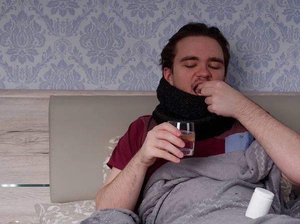 Kırmızı tişörtlü genç adam yatağa oturur ve öksürük hapları, ağrı kesiciler ve soğuk algınlığı ilaçları alır.