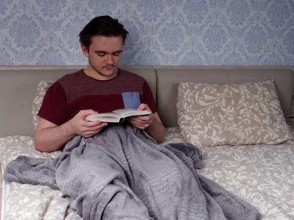 Kırmızı tişörtlü genç bir adam açık renkli bir yatakta oturur ve kalın bir kitap okur. Çalışma gününden sonra dinlenme konsepti.
