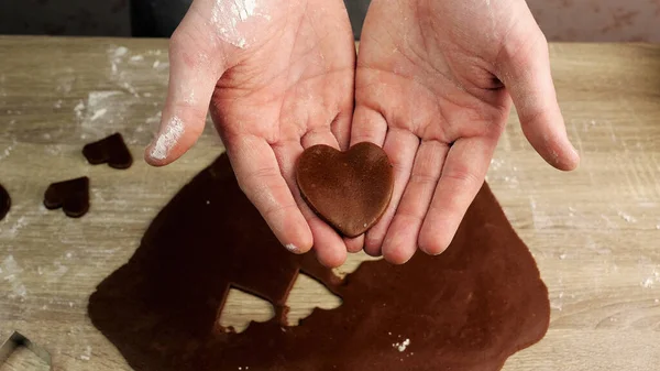 A young chef holds a heart made of ginger dough in his hand, dough and other forms in the background. Baking concept