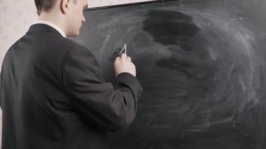 Siyah takım elbiseli bir öğretmen kara tahtaya tebeşirle, matematiksel denklemlerle yazar. Okul kavramı