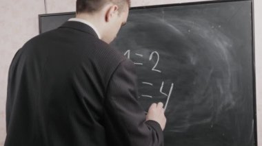 Siyah takım elbiseli bir öğretmen kara tahtaya tebeşirle, matematiksel denklemlerle yazar. Okul kavramı