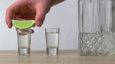 Tahta bir masanın üzerinde bir sürahi alkol var, ve yanında iki dolu bardak var, ve bir adamın eli üzerine bir limon koyuyor. Tatil ve alkol kavramı