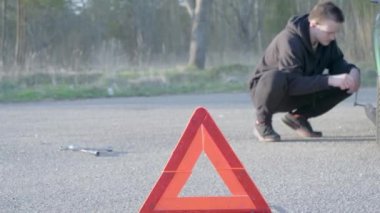 Genç bir adam kriko koyar ve arabayı kaldırır, siyah bir eşofman ve siyah bir spor ayakkabı giyer, yolda bozuk bir araba konsepti.