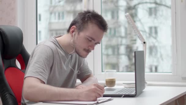 灰色のTシャツを着てコンピューターの前に座り窓のそばの白いテーブルに座り家で勉強と仕事の概念オフィスワーク — ストック動画