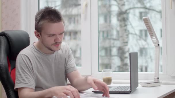 書類を読んでいる若い男がグレーのTシャツを着ていて 彼はコンピュータに座っていて 窓のそばの白いテーブルに座っていて 家で勉強と仕事の概念 オフィスワーク — ストック動画