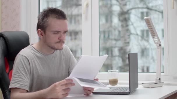 書類を読んでいる若い男がグレーのTシャツを着ていて 彼はコンピュータに座っていて 窓のそばの白いテーブルに座っていて 家で勉強と仕事の概念 オフィスワーク — ストック動画