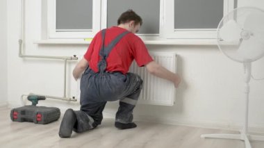 Tesisatçı odadaki merkezi ısıtma borularına beyaz metal bir radyatör yerleştiriyor. Beyaz duvarlar ve pencereler var. İnşaat ve ısıtma bakımı kavramı var.