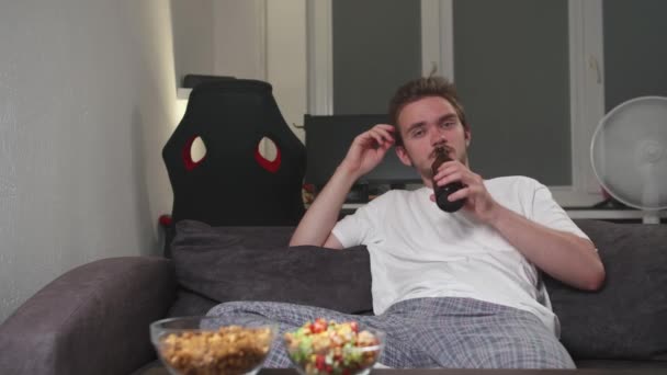 若いハンサムな髭の男がテレビを見ている 若い男がリビングルームで休んでいるとビデオを見て エンターテイメントの概念 レクリエーション ビデオゲーム テレビ ポップコーンとビール — ストック動画