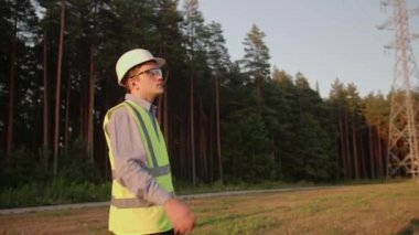 Genç elektrik mühendisi çalışmaktan yoruldu, kaskını çıkardı ve yürüyüp gitti, koruyucu giysiler ve bir şapka giyiyordu. Kavram. teknoloji, yeşil, elektrik, kule