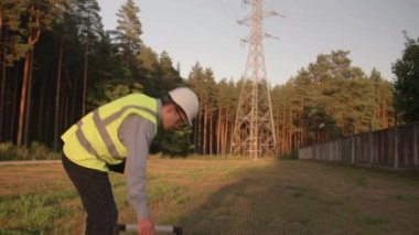 Genç bir elektrik mühendisi elektrik kulesine doğru yürür ve etrafına bakar koruyucu giysiler ve sert bir şapka giyer. Kavram. teknoloji, yeşil, elektrik, kule