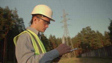 Genç bir elektrik mühendisi koruyucu giysileri ve kaskı olan bir elektrik kulesine bakıyor. Kavram. teknoloji, yeşil, elektrik, kule