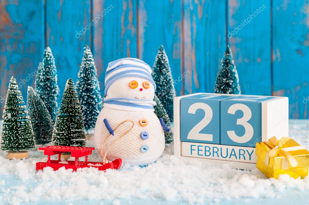 cube calendar for february 23 on wooden surface with snowman, sled, snow and fir