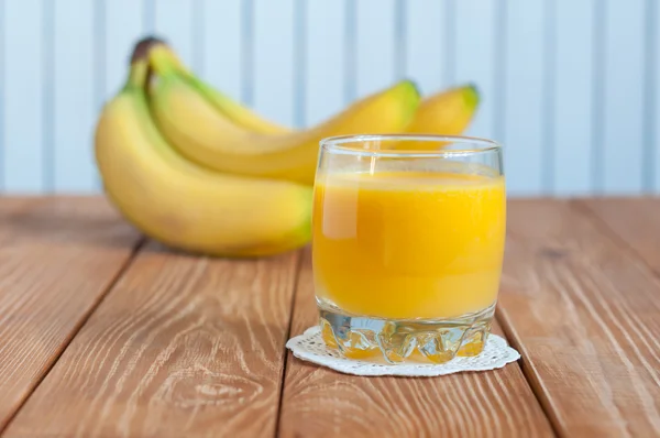 Un vaso lleno de zumo de naranja y un montón de plátanos sobre fondo de madera blanca —  Fotos de Stock