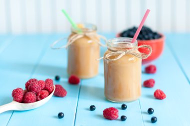 Fresh rastberry, blueberry and banana juice in jam jar on light blue wooden table, apple protein cocktails with fresh fruits.