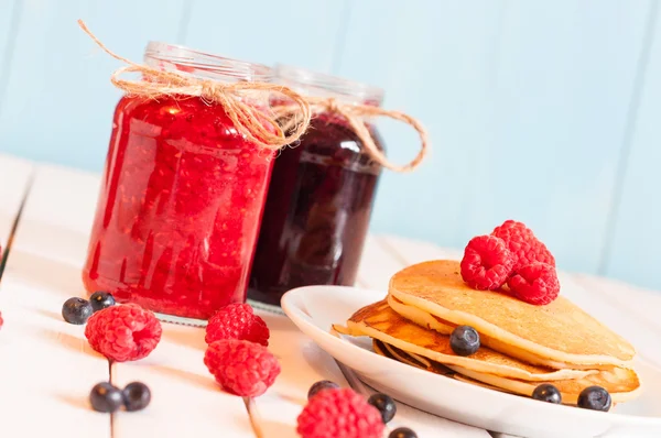 Stack of wheat golden pancakes or pancake cake with freshly picked raspberries on a dessert plate, glass mason jar full off blueberry and raspberry jam.  Unique perspective, selective focus