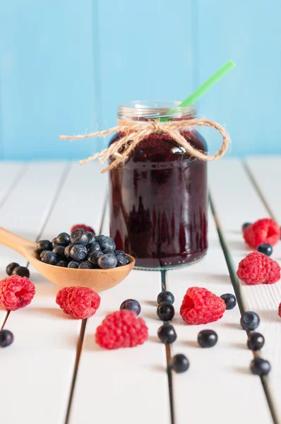 Bilberries in wood spoon. Crimson raspberry and jar of bilberry jam jn light wooden background. Selective focus — Stock Photo, Image