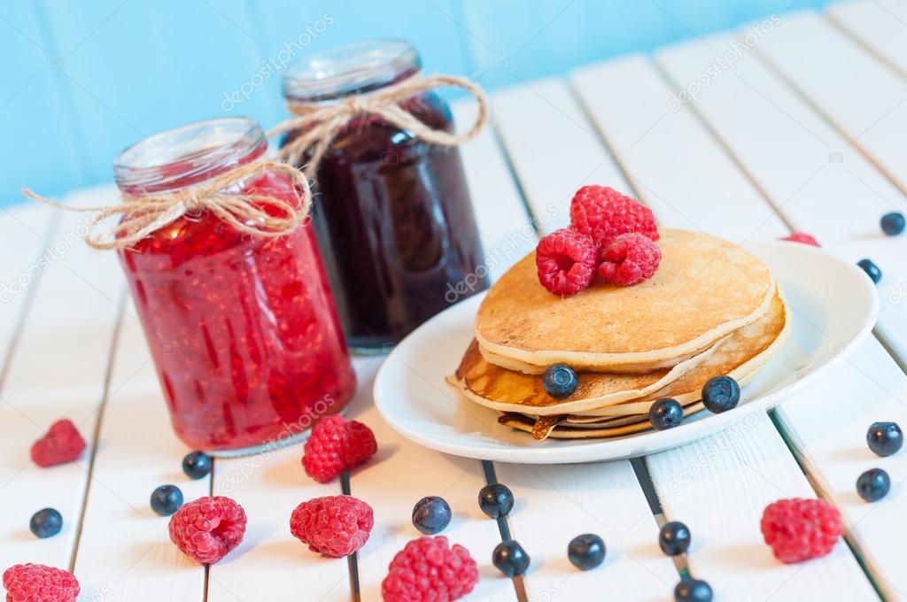 Stack of wheat golden pancakes or pancake cake with freshly picked raspberries on a dessert plate, glass mason jar full off blueberry and raspberry jam.  Selective focus, unique perspective