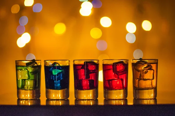 Five shots glasses full of assorted beverages and ice on golden blurred lighting bokeh background — Stock Photo, Image