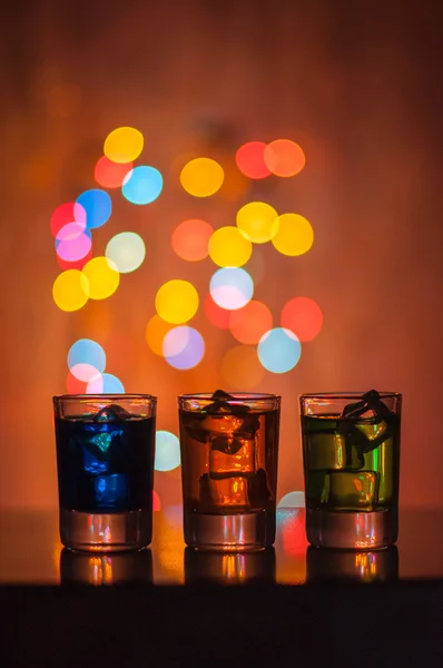 Three shots glasses full of assorted beverages and ice on golden blurred lighting bokeh background — Stock Photo, Image
