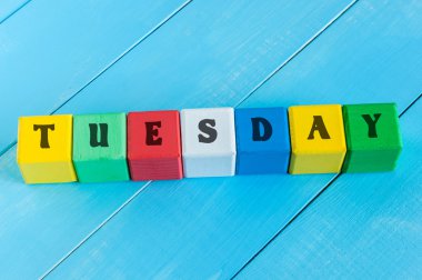 The word TUESDAY written in wooden color cubes on light blue wood background clipart
