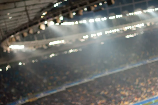 Blurred soccer stadium or ather sport arena at night with projector and stands full of people, fans, crowd. Unique perspective