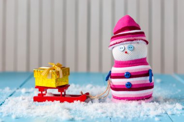 Handmade decorative snowman with red sled and present box on light wooden background. With empty space for text