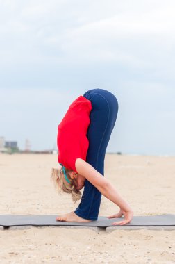 Sportif kız başı, desteklenen yapıyor yoga asana Uttanasana sahil arka plan üzerinde